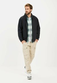 Zwarte zip-up jack met twee voorzakken over een groen geruite shirt, gecombineerd met beige cargo broeken. Witte sneakers met zwarte accenten.
