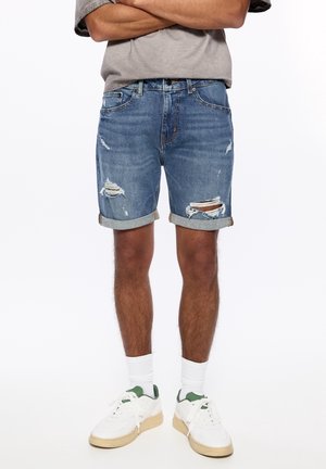 Personne portant un short en jean bleu déchiré retroussé, des chaussettes blanches et des baskets blanches, debout les bras croisés au-dessus de la taille.
