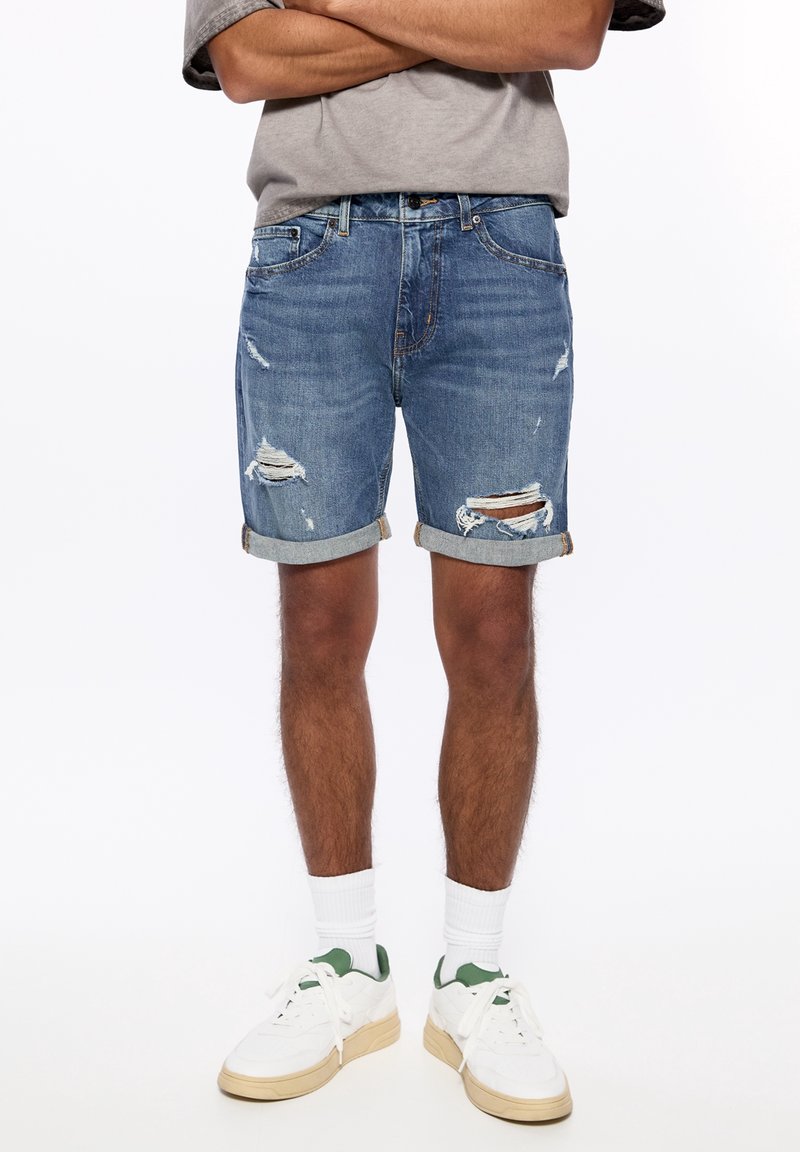 Personne portant un short en jean bleu déchiré retroussé, des chaussettes blanches et des baskets blanches, debout les bras croisés au-dessus de la taille.