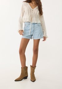 Valkoinen, pitsireunainen pitkähihaisen paita, jossa V-pääntie ja edessä napit, yhdistetty vaaleansinisiin denimshortseihin ja ruskeisiin mokkanahkaisiin nilkkureihin.