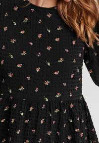 Schwarzes, texturiertes Kleid mit einem taillierten Oberteil und einem ausladenden Rock, verziert mit kleinen Blumenmustern in Rosa und Weiß. Lange Ärmel sind enthalten.