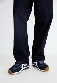 Baskets de sport bleu marine avec détails blancs, tissu texturé et semelle en caoutchouc, associées à un pantalon en denim foncé avec coutures apparentes.