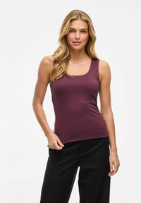 Donna con capelli lunghi biondi che indossa un top senza maniche di colore prugna e pantaloni neri, in piedi contro uno sfondo bianco semplice.
