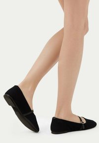 Svart tygskor med rund tå och rem över fotryggen. Strukturerad yta och låg klack med en diskret detalj nära remmen.