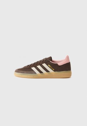 Brązowy but sportowy Adidas Spezial z białymi paskami, różowym kołnierzem przy pięcie, gumową podeszwą i złotym logo po boku, pokazany z profilu.