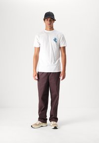 Hvid bomulds-T-shirt med lille blå håndgrafik, parret med mørk maroon bukser og beige sneakers med sorte accenter. Sort kasket på hovedet.