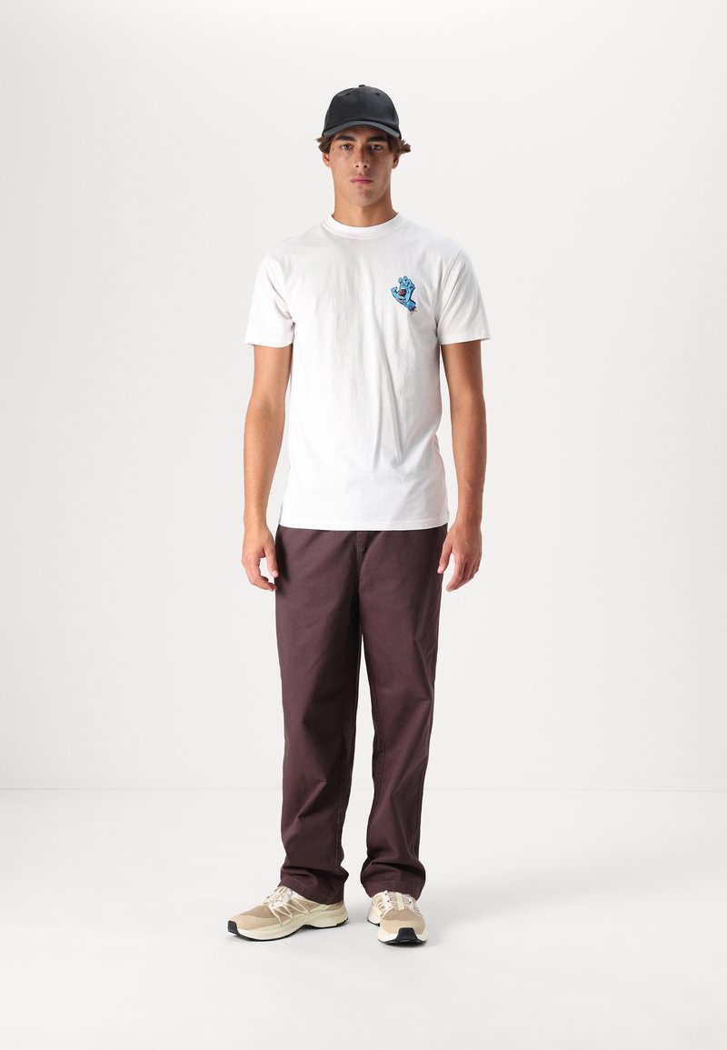 Hvid bomulds-T-shirt med lille blå håndgrafik, parret med mørk maroon bukser og beige sneakers med sorte accenter. Sort kasket på hovedet.