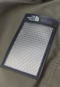 North Face produktdetalj med en rektangulær, strukturert grålapp med sekssidig mønster, omgitt av en mørkeblå kant og logo.