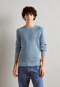 Hellblauer gestrickter Pullover mit einem strukturierten Muster, gerippten Bündchen und Saum. Kombiniert mit blauen Jeans und einer schlichten Kettenhalskette.