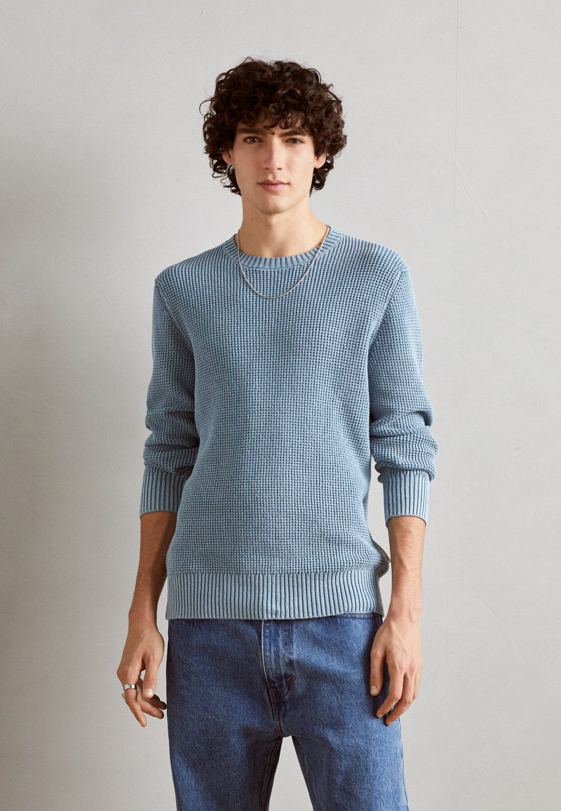 Hellblauer gestrickter Pullover mit einem strukturierten Muster, gerippten Bündchen und Saum. Kombiniert mit blauen Jeans und einer schlichten Kettenhalskette.