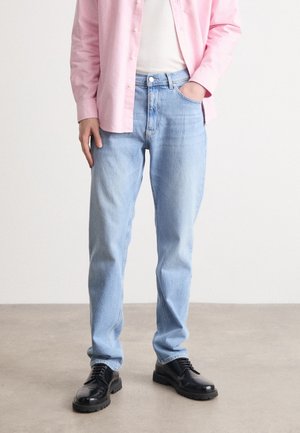 Osoba ubrana w jasnoniebieskie dżinsy, czarne sznurowane buty, białą koszulę oraz różową koszulę zapinaną na guziki, z jedną ręką w kieszeni.