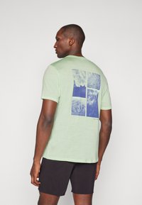 T-shirt verde claro de mangas curtas com um design gráfico azul de quatro imagens nas costas, com decote redondo e textura suave.