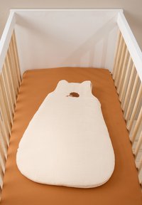 Baumwoll-Schlafsack in Creme mit hellen Streifen, runder Form und kleiner, brauner Igel-Stickerei. Auf einer terrakottafarbigen Matratze platziert.