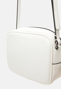 Biały torebka crossbody z sztucznej skóry o prostokątnym kształcie, z zamkiem błyskawicznym, wytłoczonym logo z przodu oraz czarnymi akcentami metalowymi.