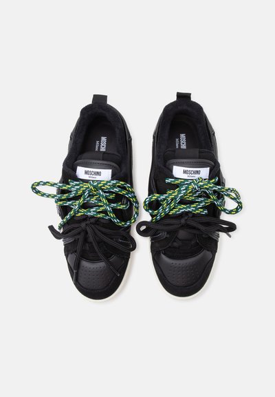 Ζευγάρι μαύρα sneakers Moschino με χοντρές πράσινες και κίτρινες κορδόνια με σχέδιο πάνω από τα συνηθισμένα μαύρα κορδόνια, από πάνω.