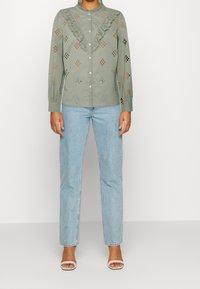 Olijfgroene shirt met een ruches kraag, kanten details en diamanten uitsparingen. Gecombineerd met lichtblauwe rechte jeans en witte sandalen.