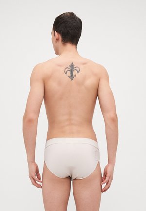 Beige Slip mit glatter Textur und einem breiten Bund, mit einem Logo an der Seite und einem Fleur-de-Lis-Tattoo auf der Rückseite.