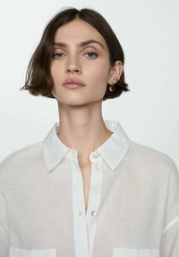 Camicia bianca con colletto, realizzata in un tessuto leggero e leggermente trasparente, dotata di taschino frontale e bottoni perla.