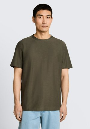 Man met kort zwart haar die een effen olijfgroen T-shirt met korte mouwen en lichtblauwe jeans draagt, staand tegen een lichte achtergrond.