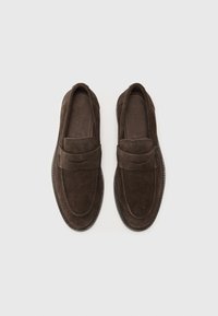 Mocasines de ante marrón con un diseño de mocasín, que presentan una correa frontal, detalles de costura y suelas de goma. Estilo plano y tipo slip-on.