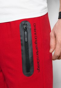 Un pantalon de survêtement rouge présente une poche zippée noire sur le côté, avec le texte "Ombre Original Men's Design" imprimé en noir le long de la jambe.