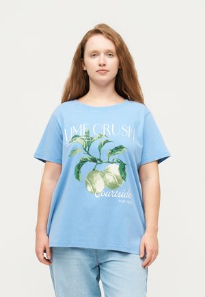 Ung kvinde med langt rødt hår iført en blå T-shirt med grafik af lime og blad samt lyseblå jeans, stående foran en ensfarvet baggrund.