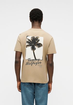 Uomo con maglietta beige con grande grafica di palma e testo "Tommy Hilfiger" sul retro, abbinata a jeans blu, in piedi di spalle.