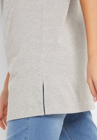 Maglietta grigia in morbido cotone con uno spacco laterale. Il tessuto ha una consistenza liscia e la maglietta è abbinata a pantaloni jeans blu.