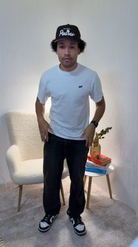 Wit T-shirt met een klein zwart logo, donkere spijkerbroek, zwart-witte sneakers, zwarte pet met de tekst "San Diego Padres", staand in een kamer met meubels.