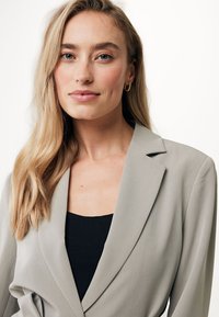 Blazer grigio chiaro con un design strutturato, colletto a risvolto e un nastro in vita, indossato su una maglietta nera. Tessuto liscio e leggero.