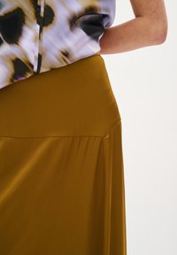Jupe fluide couleur moutarde avec une texture lisse, taille haute et plis subtils. Associée à un chemisier à motifs avec des couleurs douces et des formes abstraites.