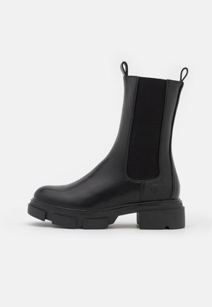 Bota Chelsea de cuero negro con suela gruesa de tacos, paneles laterales elásticos y tiradores en la parte delantera y trasera sobre fondo liso y claro.