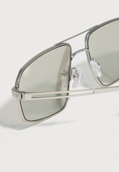 Ασημένια μεταλλικά γυαλιά ηλίου τύπου πιλότου με γκρι φακούς και διαφανή ρυθμιζόμενα μαξιλαράκια μύτης πάνω σε λευκή επιφάνεια.