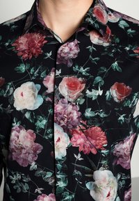 Blommigt mönstrad skorta med mörk bakgrund och stora rosor i rosa och vitt, accentuerad med gröna blad. Knapp-up design med klassisk krage.