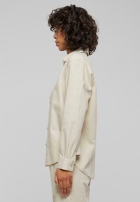 Beige skjorte med knapper og en avslappet passform, med lange ermer, krage, sideslisser og en jevn tekstur.