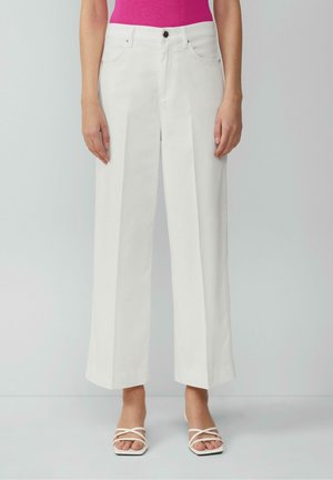 Femme portant un pantalon blanc taille haute à jambes larges avec un pli sur le devant, associé à un haut rose et des talons blancs ouverts à brides, debout devant un fond uni.