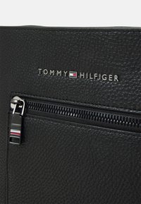 Borsa in pelle nera a trama con tasca con zip, hardware argentato e logo "TOMMY HILFIGER" impresso con accenti colorati.