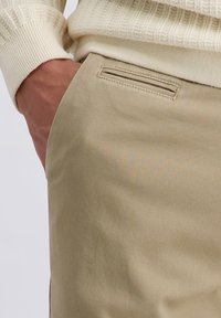 Männerhand in der Tasche einer beigen Hose, trägt einen cremefarbenen Strickpullover, Fokus auf den Taschendetails und dem strukturierten Stoff.