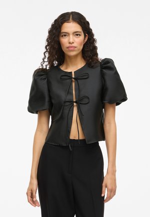 Blusa nera corta con maniche a sbuffo, texture satinata, chiusura con fiocco frontale e apertura a chiave. Abbinata a pantaloni neri.