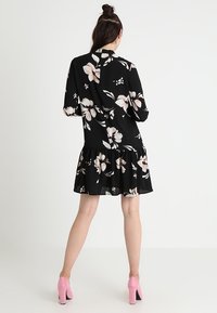 Robe florale noire à manches longues, avec un ourlet volanté et un col rond. Présente de grands motifs de fleurs roses et blanches. Associée à des escarpins roses.