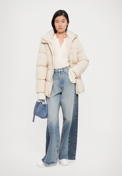 Giubbotto softshell beige, maglione a coste bianco, jeans a gamba larga con sfumature blu, sneakers bianche e borsa blu con dettagli in argento.