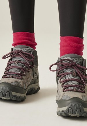 Jambes en collants noirs, portant des chaussures de randonnée grises et noires avec des lacets roses et des chaussettes roses vives froncées, debout sur un fond neutre.