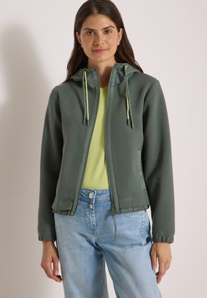 Frau trägt eine grüne Zip-up-Kapuzenjacke über einem hellgrünen Oberteil und hellblauen hoch taillierten Jeans, stehend vor einem einfarbigen Hintergrund.