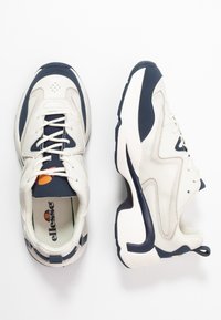 Sneakers bianchi e navy con tomaia in pelle texturizzata, dettagli perforati e lacci a contrasto. Presentano una suola spessa e un marchio sulla linguetta.