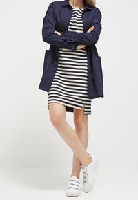 Giacca navy a maniche lunghe sopra un vestito a righe bianco e nero con spacco laterale, abbinata a sneaker bianche con tre cinturini.