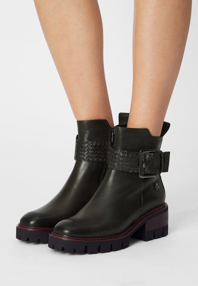 Bottines en cuir noir avec talons épais, lanière tressée et détail à boucle, portées sur des jambes nues sur un fond clair uni.