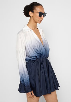Donna che indossa un vestito lungo a maniche lunghe blu sfumato, con scollatura a V profonda e gonna svasata, capelli raccolti a chignon e occhiali da sole neri, di profilo.
