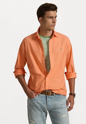 Νεαρός άντρας που φοράει ένα ανοιχτό πορτοκαλί πουκάμισο με κουμπιά πάνω από ένα πράσινο και άσπρο ριγέ μπλουζάκι, με γυρισμένα μανίκια, ανοιχτό μπλε τζιν και καφέ ζώνη.