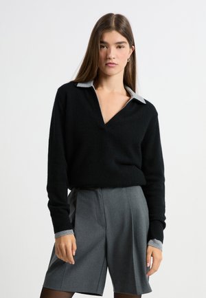 Schwarzer, V-Ausschnitt Pullover aus weichem Material mit grauem Kragen und Bündchen. Kombiniert mit grauen, taillierten Shorts und strukturierten Strumpfhosen.