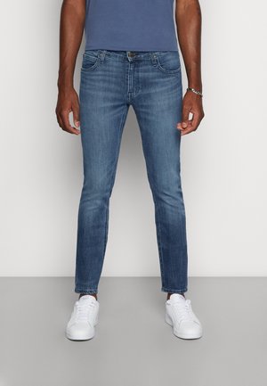 Mann trägt ausgewaschene blaue Slim-Fit-Jeans, weiße Turnschuhe, ein blaues Hemd und ein silbernes Armband, steht vor einer schlichten Wand.